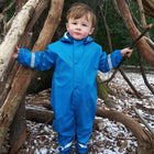 A young boy in a blue Rainy Day Waterproof Puddle Suit stands amongst snowy leaves and branches in the woods.
